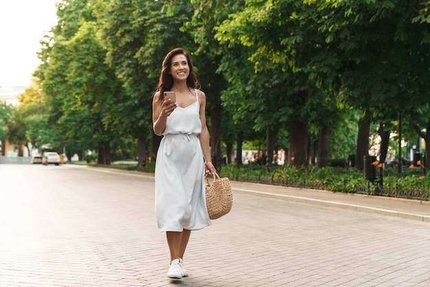 Portret Szczęśliwej Kobiety W Letniej Sukience Uśmiecha Się I Trzyma Telefon Podczas Spaceru Po Zielonym Bulwarze