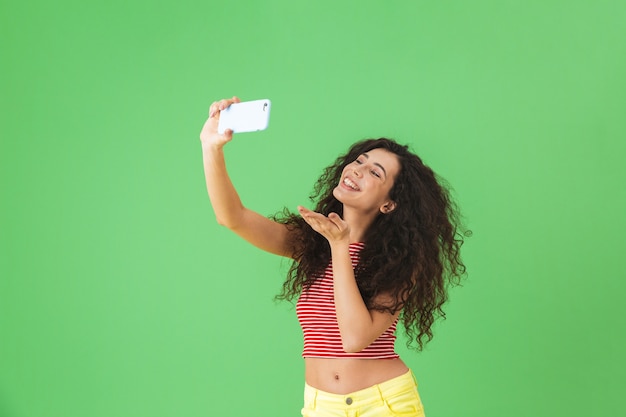 Portret szczęśliwej kobiety w wieku 20 lat w letnich ubraniach, uśmiechniętej i używającej telefonu komórkowego do selfie na zielono