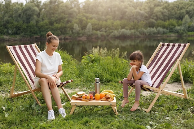 Portret Szczęśliwej Matki Z Córką Siedzącą Na Składanych Krzesłach Piknikowych I Jedzących Owoce I Kanapki Podczas Pikniku W Pobliżu Rzeki, Ciesząc Się Spędzaniem Czasu Razem I Piękną Przyrodą