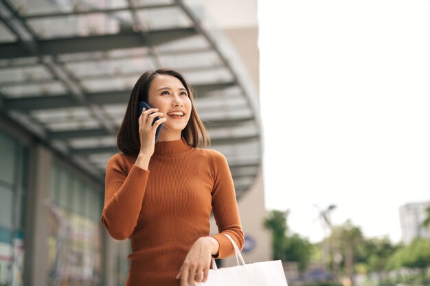 Portret szczęśliwej młodej azjatyckiej kobiety rozmawiającej przez telefon komórkowy i niosącej papierowe torby na zakupy spacerujące po centrum handlowym na świeżym powietrzu