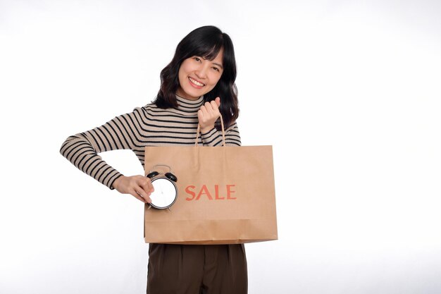 Portret szczęśliwej młodej azjatyckiej kobiety z koszulą ze swetrem trzymającej budzik i papier na zakupy z powrotem na białym tle