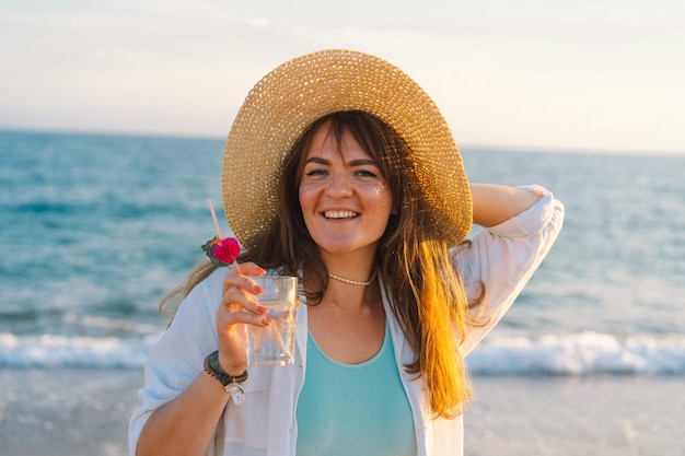 Portret szczęśliwej młodej dziewczyny w słomkowym kapeluszu z koktajlem w ręku na tle morza