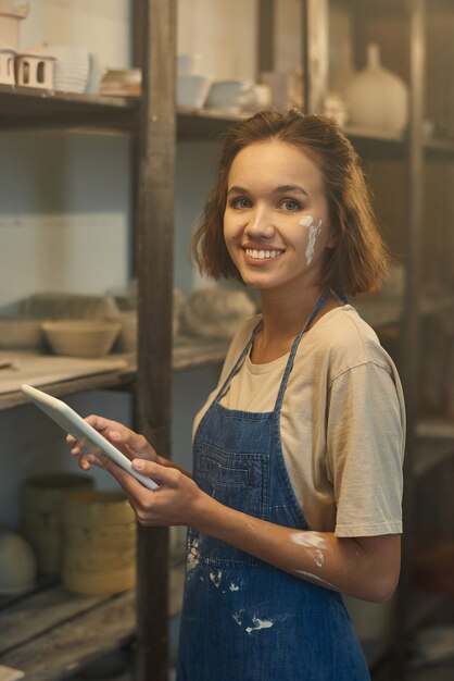 Portret szczęśliwej młodej garncarki w dżinsowym fartuchu przy użyciu cyfrowego tabletu w magazynie ceramiki