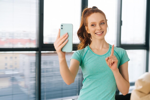 Portret szczęśliwej młodej kobiety w przypadkowych ubraniach rozmawiającej przez rozmowę wideo, trzymającej w ręku smartfon, strzelającej do selfie przednią kamerą i pokazującej palec wskazujący w górę