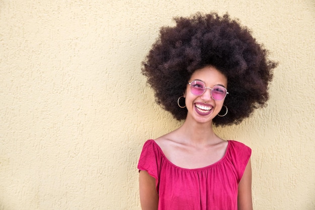 Portret szczęśliwej młodej marokańskiej kobiety z fryzurą afro i stylowymi okularami przeciwsłonecznymi stojącej w pobliżu beżowej ściany i patrzącej na kamerę