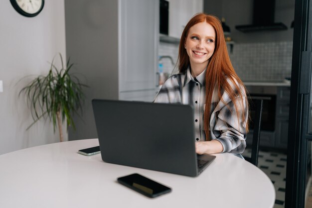 Portret szczęśliwej młodej rudowłosej kobiety siedzącej przy biurku z laptopem i uśmiechniętym telefonem komórkowym odwracającym wzrok