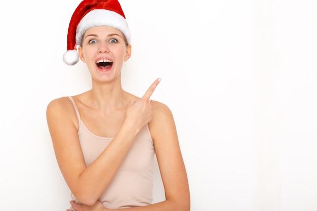 Portret szczęśliwej modelki w czerwonym kapeluszu noworocznym wskazuje palcem wskazującym miejsce na reklamę lub tekst promocyjny. Śliczna dziewczynka.