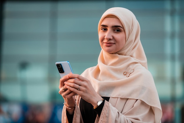 Portret szczęśliwej muzułmańskiej bizneswoman w hidżabie używającej telefonu w pobliżu budynku biurowego
