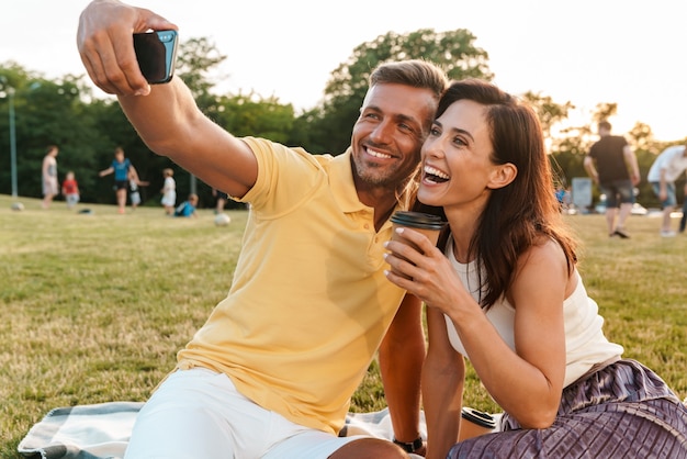 Portret szczęśliwej pary dorosłych mężczyzny i kobiety pije kawę na wynos i robi zdjęcie selfie na telefonie komórkowym, siedząc na trawie w parku