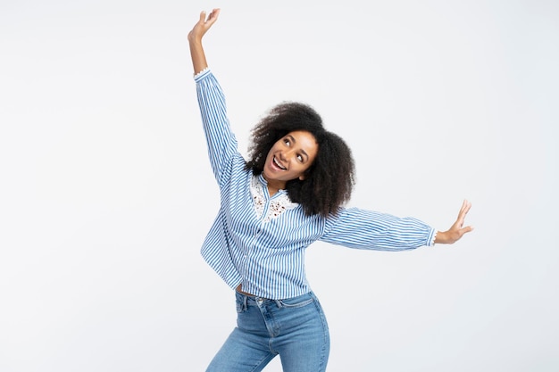 Portret szczęśliwej, pięknej, autentycznej Afroamerykanki z kręconymi włosami, ubranej w luźną koszulę, odwracającej wzrok od rąk na bok, gest latania izolowany na białym tle