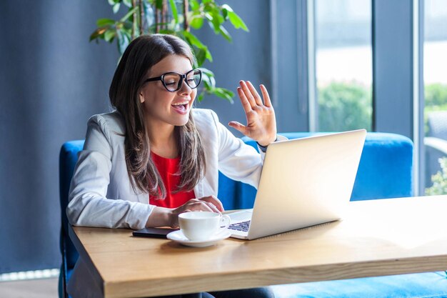 Portret szczęśliwej pięknej stylowej brunetki młodej kobiety w okularach siedzącej, patrząc na ekran jej laptopa na rozmowie wideo i pozdrowienie ze zdziwioną twarzą kryty strzał studio kawiarnia tło biurowe