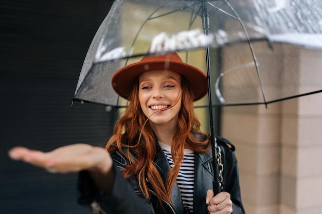 Portret szczęśliwej rudowłosej młodej kobiety w modnym kapeluszu podnoszącej rękę do sprawdzenia, czy deszcz przestanie próbować złapać spadające krople deszczu