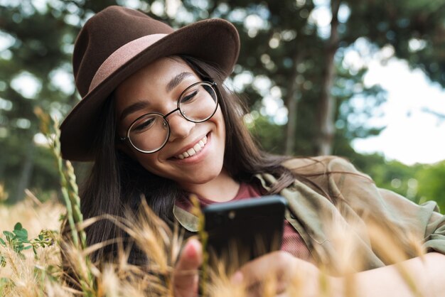 Portret Szczęśliwej Stylowej Kobiety O Długich Ciemnych Włosach, W Kapeluszu I Okularach, Korzystającej Z Telefonu Komórkowego, Leżąc Na Trawie W Zielonym Parku