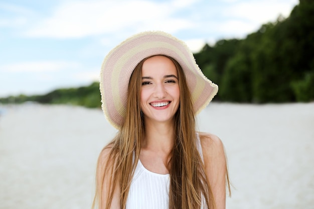 Portret szczęśliwej uśmiechniętej kobiety w błogości wolnego szczęścia na plaży oceanu stojącej w kapeluszu. Modelka w białej letniej sukience ciesząca się przyrodą podczas wakacji na świeżym powietrzu