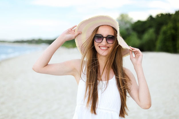 Portret szczęśliwej uśmiechniętej kobiety w wolnym szczęściu na plaży oceanu, stojącej w kapeluszu i okularach przeciwsłonecznych. Modelka w białej letniej sukience ciesząca się przyrodą podczas wakacji w podróży
