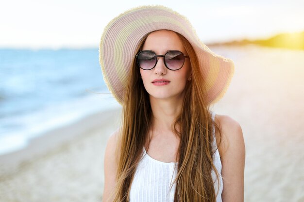 Portret szczęśliwej uśmiechniętej kobiety w wolnym szczęściu na plaży oceanu, stojącej w kapeluszu i okularach przeciwsłonecznych Modelka w białej letniej sukience ciesząca się przyrodą podczas wakacji w podróży