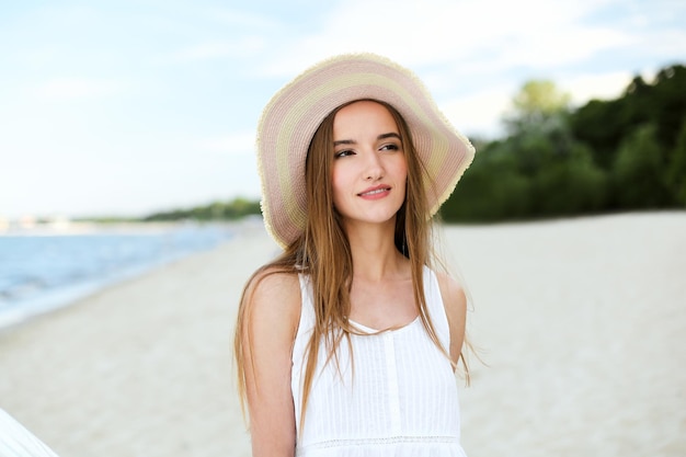 Portret szczęśliwej uśmiechniętej kobiety w wolnym szczęściu na plaży oceanu, stojącej w kapeluszu. Modelka w białej letniej sukience cieszy się przyrodą podczas wakacji na świeżym powietrzu.