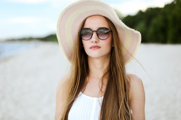 Portret szczęśliwej uśmiechniętej kobiety w wolnym szczęściu na plaży oceanu stojącej z kapeluszem i okularami przeciwsłonecznymi. Kobieta model w białej letniej sukience cieszy się przyrodą podczas wakacji