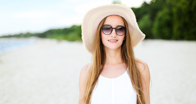 Portret szczęśliwej uśmiechniętej kobiety w wolnym szczęściu na plaży oceanu stojącej z kapeluszem i okularami przeciwsłonecznymi.