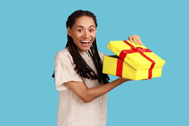 Portret szczęśliwej zdumionej kobiety z czarnymi dredami, patrzącej do pudełka prezentowego, otwierającej prezent i patrzącej ze szczęściem na kamerę, ubrana w białą koszulę. Kryty studio strzał na białym tle na niebieskim tle.