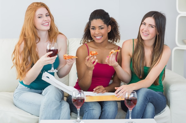 Portret szczęśliwi młodzi żeńscy przyjaciele z pizzą i winem na kanapie w domu