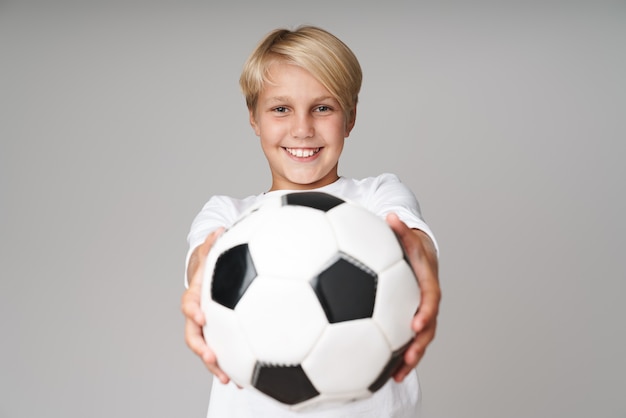 Portret Szczęśliwy Blond Chłopiec Pozowanie Na Białym Tle Nad Szarą ścianą Trzymając Piłkę Nożną.