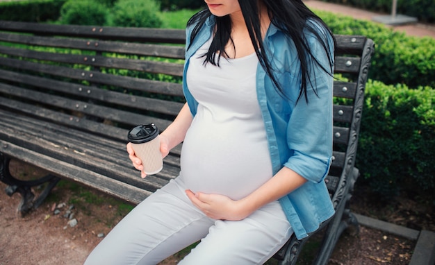 Portret szczęśliwy czarne włosy i dumny kobieta w ciąży w parku. Modelka siedzi na miejskiej ławce i pije kawę lub herbatę.