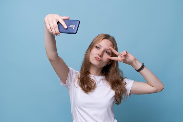 Portret Szczęśliwy Przyjazny Młody Kaukaski Dziewczyna W Białej Koszulce Na Niebieskim Tle Biorąc Selfie I Macha Ręką, Wideorozmowa, Czat Online. Pojęcie Komunikacji Przez Internet