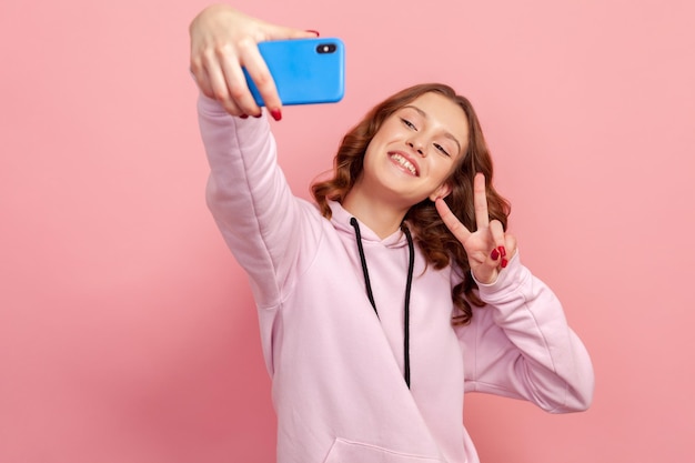 Portret Szczęśliwy Teen Dziewczyna W Bluzie Z Kapturem Pokazując Gest Zwycięstwa Palcami Na Aparacie Smartfona I Szczerze Uśmiechnięty Vlog Lub Selfie Kryty Studio Strzał Na Białym Tle Na Różowym Tle