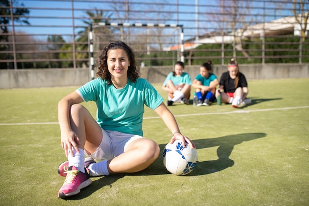 Zdjęcie portret szczęśliwy żeński piłkarz pozowanie na polu. piękna dziewczyna siedzi z piłką patrząc na kamery i inne dziewczyny odpoczynek z tyłu, komunikując się. hobby, rekreacja, koncepcja sportów zespołowych