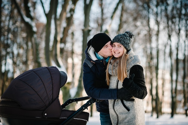 Portret szczęśliwych młodych rodziców stoją i całują się z dzieckiem wózka w winter park