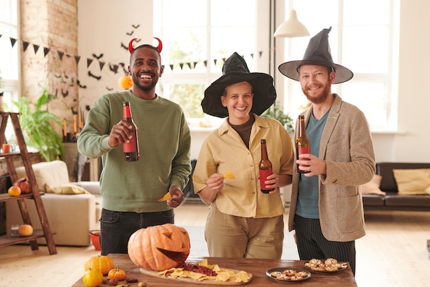 Portret szczęśliwych młodych wieloetnicznych przyjaciół w halloweenowych opaskach stojących z nachos i piwnym bot