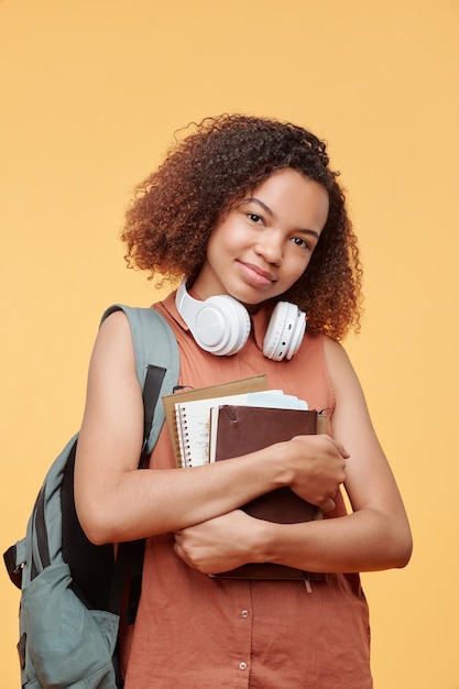 Portret treści piękny afroamerykański uczeń szkoły średniej z bezprzewodowymi słuchawkami na szyi obejmujący skoroszyty na żółtym tle
