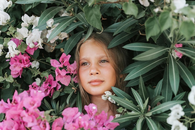 Portret twarz szczerej pięknej małej dziewczynki w wieku ośmiu lat z zielonymi brązowymi oczami na tle zielonych roślin i różowych kwiatów podczas letnich wakacji podróży gen z koncepcja zdrowia psychicznego