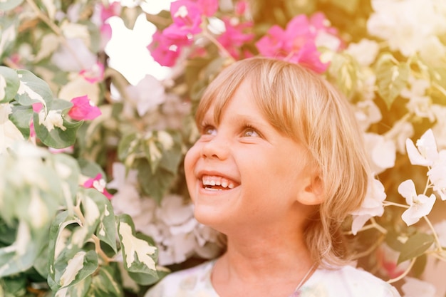 Portret twarzy szczery mały szczęśliwy uśmiechający się pięcioletni blond chłopiec z zielonymi oczami w różowo-białych roślinach kwiatowych w przyrodzie dzieci bawią się letnie wakacje jasne światło i przewiewna flara