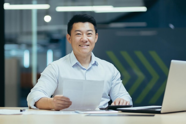 Portret udanego azjatyckiego szefa z dokumentem w nowoczesnym biurze uśmiechający się i patrzący na kamerę