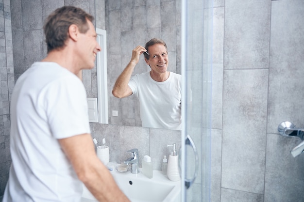 Portret uniesionego do pasa mężczyzny w dobrym humorze, czeszącego włosy przed lustrem łazienkowym