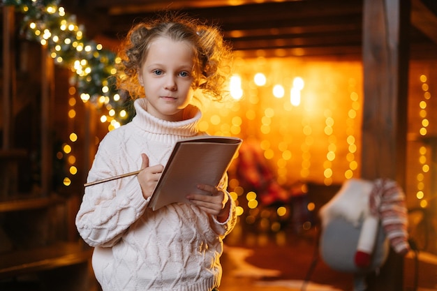Portret urocza szczęśliwa mała blondynka kręcone dziecko dziewczyna pisze list do Świętego Mikołaja patrząc na kamerę w domu z świątecznym jasnym wnętrzem