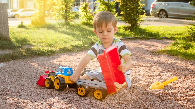 Portret uroczego 3-letniego chłopca bawiącego się zabawkową ciężarówką z przyczepą na placu zabaw w parku