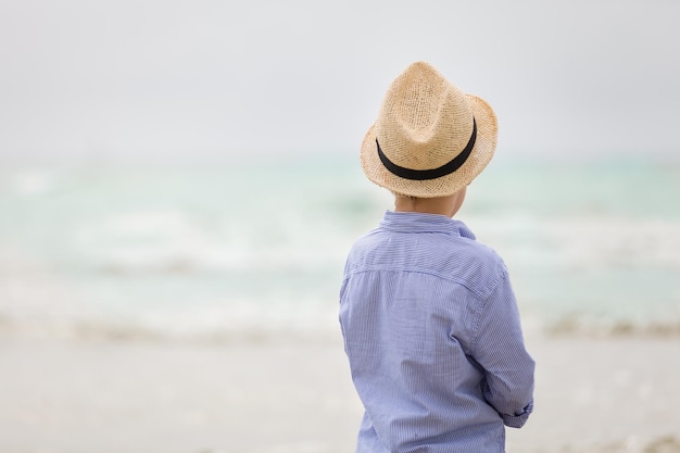 Portret uroczego chłopca w słomkowym kapeluszu spacerującego po plaży nad oceanem i patrzącego na fale