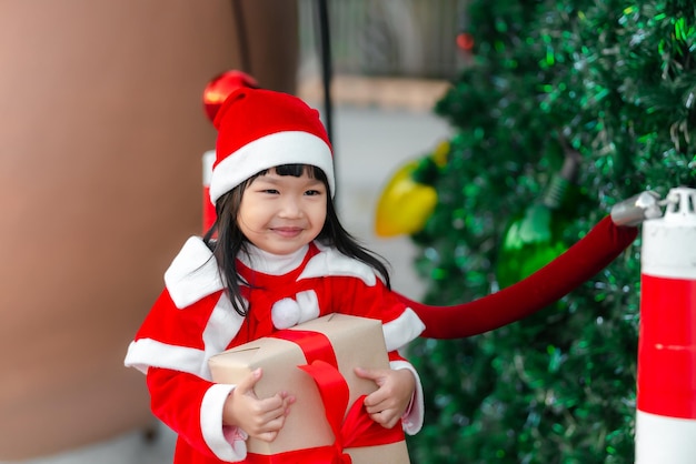 Portret uroczej azjatyckiej małej dziewczynki nosi sukienkę świętego mikołaja szczęśliwego w pobliżu choinki.Tajlandia dziecko dołącza do festiwalu świątecznego