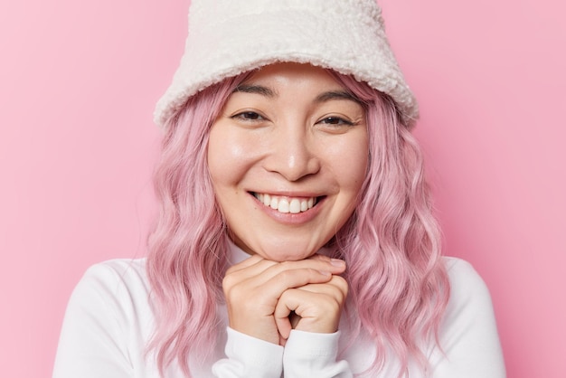 Portret uroczej azjatyckiej nastolatka trzyma ręce pod brodą, uśmiecha się szczęśliwie, wpatrując się szczęśliwie w kamerę, nosi futrzany kapelusz i biały poloneck ma ufarbowane na różowo włosy pozuje w pomieszczeniu Pozytywne ludzkie emocje