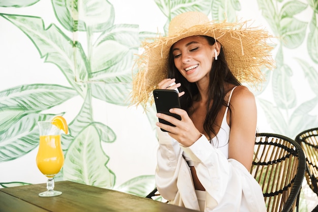 Portret uroczej brunetki w słomkowym kapeluszu i letnich ubraniach za pomocą smartfona podczas picia soku pomarańczowego w kawiarni na plaży