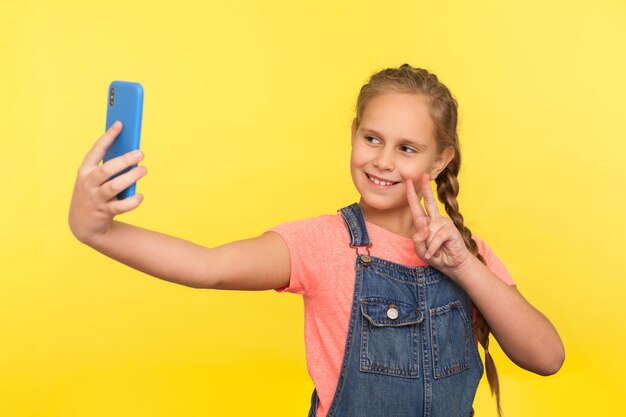 Portret uroczej dziewczynki w dżinsowym kombinezonie gestem zwycięstwa lub pokoju podczas komunikowania się z rodzicami podczas rozmowy wideo, robienia selfie na telefonie komórkowym. studio strzał na białym tle na żółtym tle