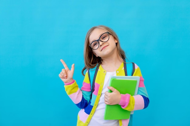 Portret uroczej dziewczynki w okularach w pasiastej kurtce z zeszytami i podręcznikami w dłoniach oraz plecakiem pokazuje kciuki do góry Koncepcja edukacji niebieskie tło miejsce na tekst