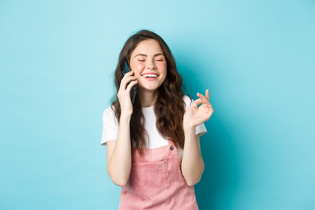 Portret uroczej i stylowej dziewczyny śmiejącej się podczas rozmowy przez telefon, trzymającej smartfon w pobliżu ucha i uśmiechniętej szczęśliwie, prowadzącej swobodną rozmowę, stojącej na niebieskim tle.