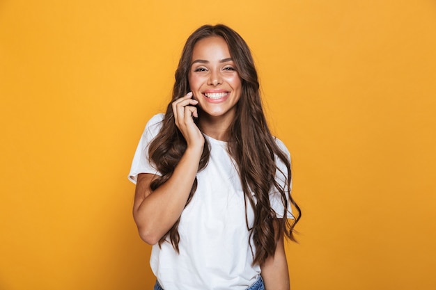 Portret uroczej kobiety 20s z długimi włosami, uśmiechając się i rozmawiając przez telefon komórkowy, na białym tle nad żółtą ścianą