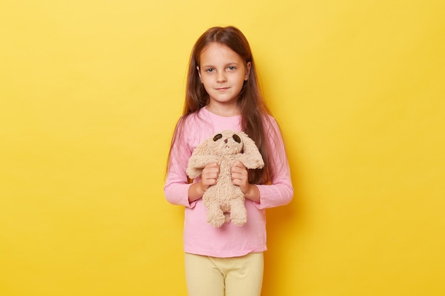 Portret uroczej małej kaukaskiej dziewczynki trzymającej puszystą zabawkę w rękach stojącej na żółtym tle patrzącej na kamerę w swobodnym ubraniu
