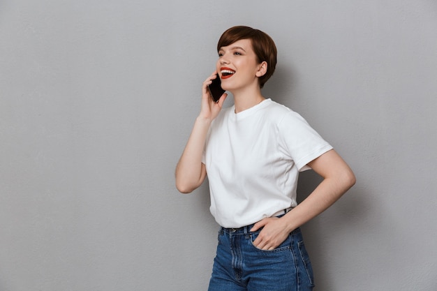 Portret uroczej młodej kobiety uśmiecha się i rozmawia przez telefon komórkowy na białym tle nad szarą ścianą
