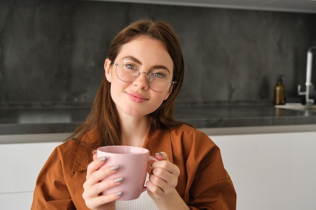Portret uroczej młodej kobiety w okularach siedzi w domu z filiżanką herbaty dziewczyna w kuchni cieszy